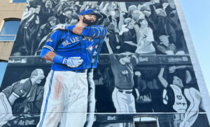 Large outdoor mural of Toronto Blue Jays baseball player Jose Bautista in a dynamic batting pose, prominently featuring the team's blue and white uniform against a grayscale crowd background, highlighting sports culture and public art in an urban setting.